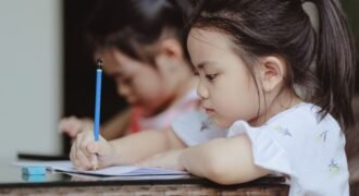 Adorable focused little Asian preschool girls sitting at table and doing tasks in copybooks in classroom